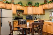 A kitchen area with wooden cabinets, a refrigerator, a microwave, a coffee maker, and a round wooden dining table with four chairs. There are decorative plants on top of the cabinets and a light green wall in the background.