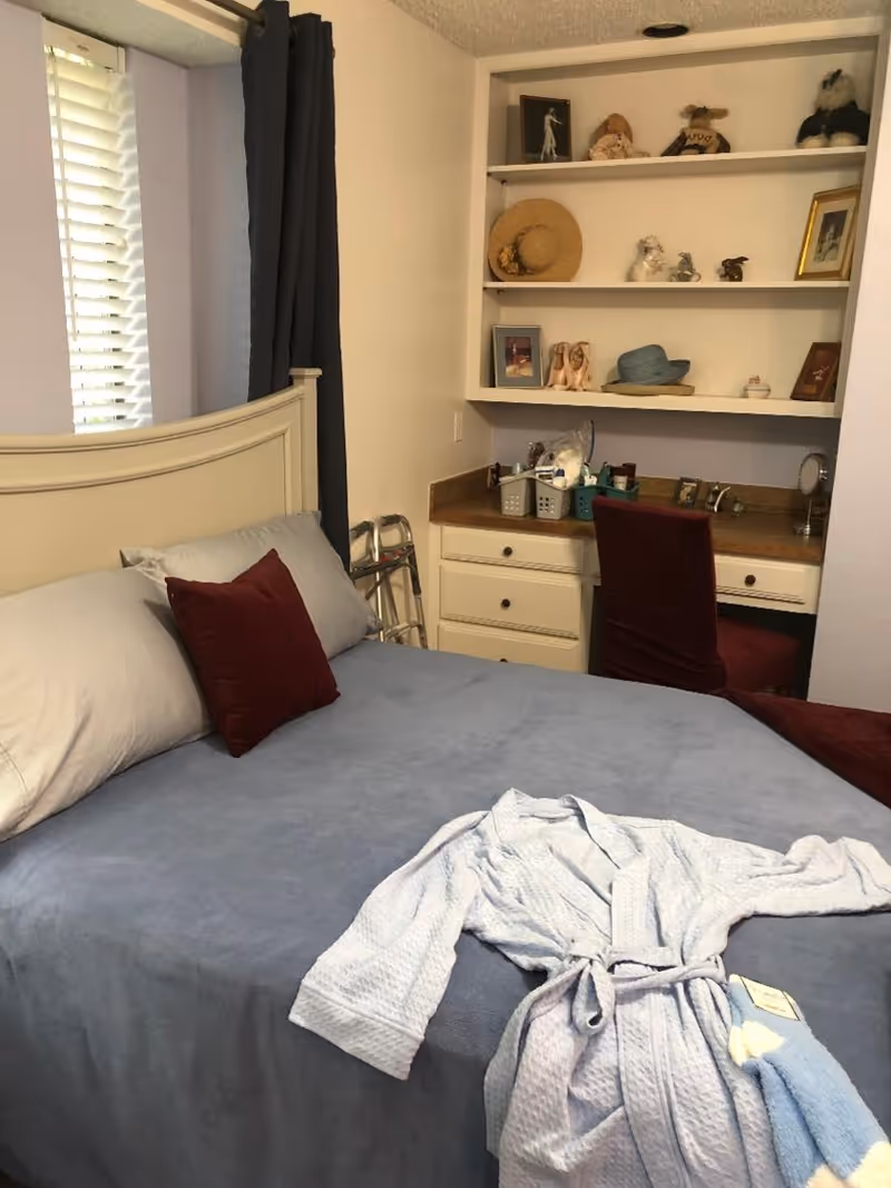A neatly made bedroom with a bed, desk, shelves, and a walker beside the bed.