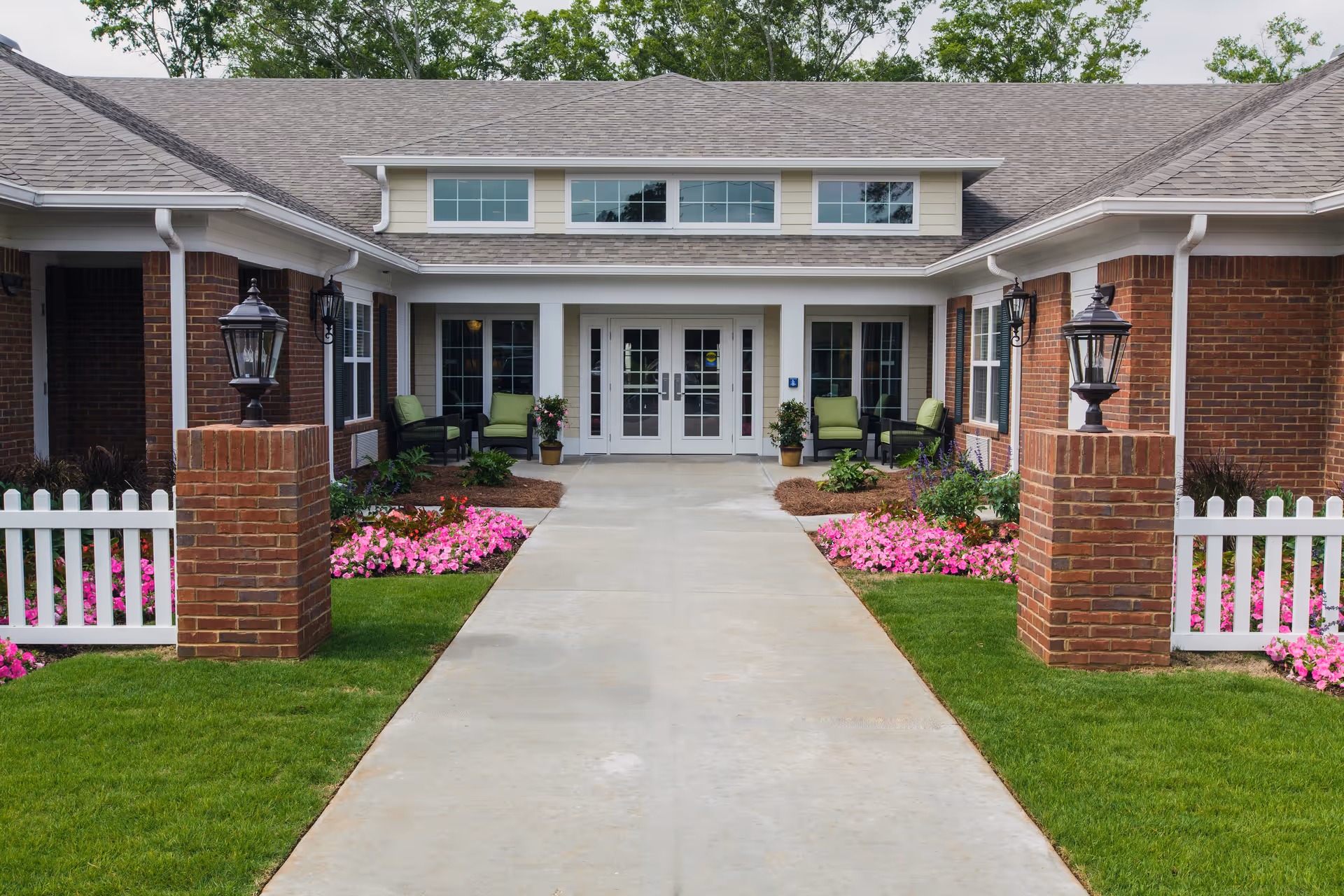 Front entrance of a senior living facility with a concrete walkway leading to double glass doors. The building has a brick exterior with white trim and a gray shingled roof. There are green cushioned chairs on either side of the entrance, surrounded by flower beds with pink flowers and green grass. Two brick pillars with black lanterns flank the walkway near the entrance.