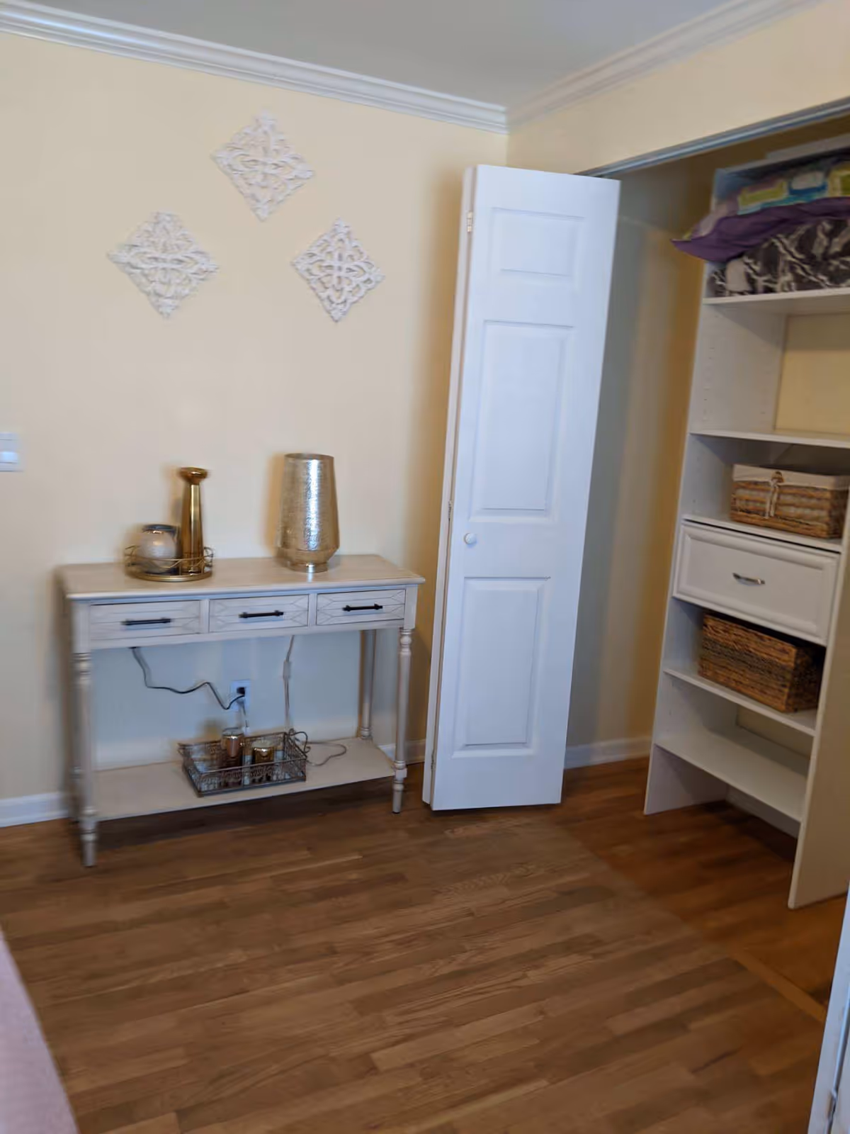 A small room with light yellow walls and wooden flooring. There is a white console table with three drawers against the wall, decorated with a gold vase, a candle holder, and a small tray with candles underneath. To the right, there is an open closet with shelves containing folded blankets and wicker baskets. Three decorative white wall hangings are arranged in a diamond pattern above the console table.