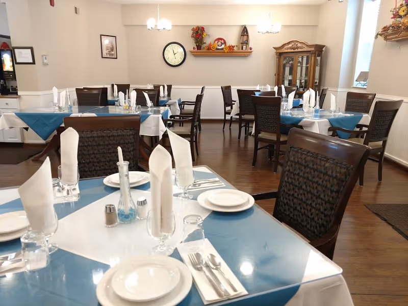 Dining room with multiple tables set with white plates, silverware, glasses, and folded white napkins. The tables have blue and white tablecloths. The room has wooden chairs, a wooden floor, a clock on the wall, a wooden cabinet, and autumn-themed decorations on a shelf and wall.