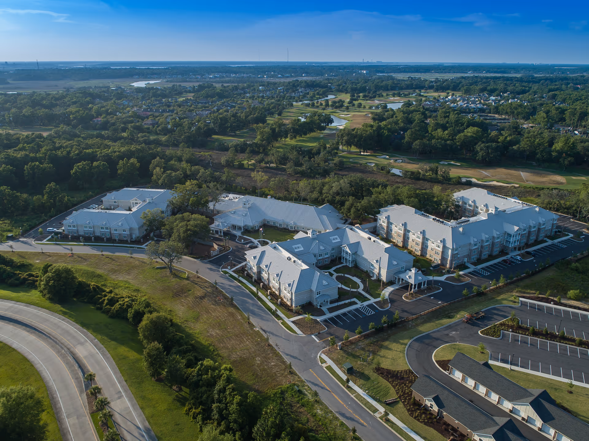 Aerial view of Wellmore of Daniel Island senior living facility surrounded by greenery, roads, and parking areas, with a golf course and residential neighborhood in the background under a clear sky.