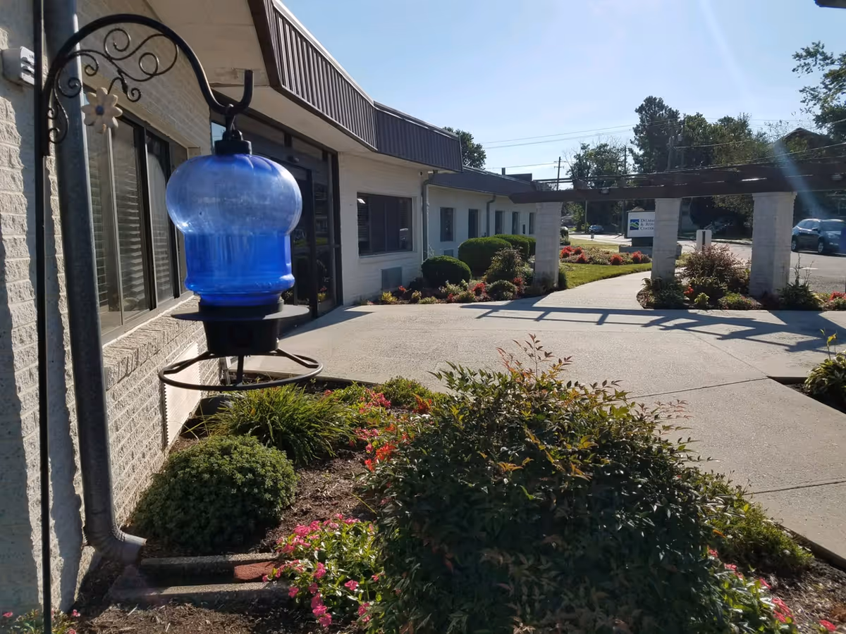 Outdoor view of a nursing home entrance with a blue glass lantern in the foreground, landscaped flower beds, a paved walkway and columns leading to the building.