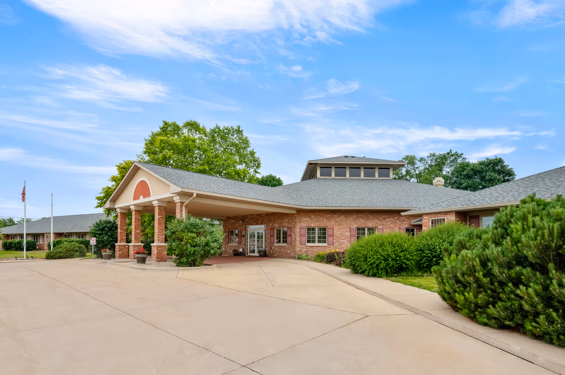 Front entrance of a single-story brick senior living facility with a covered porte-cochere, driveway, and landscaping under a blue sky.