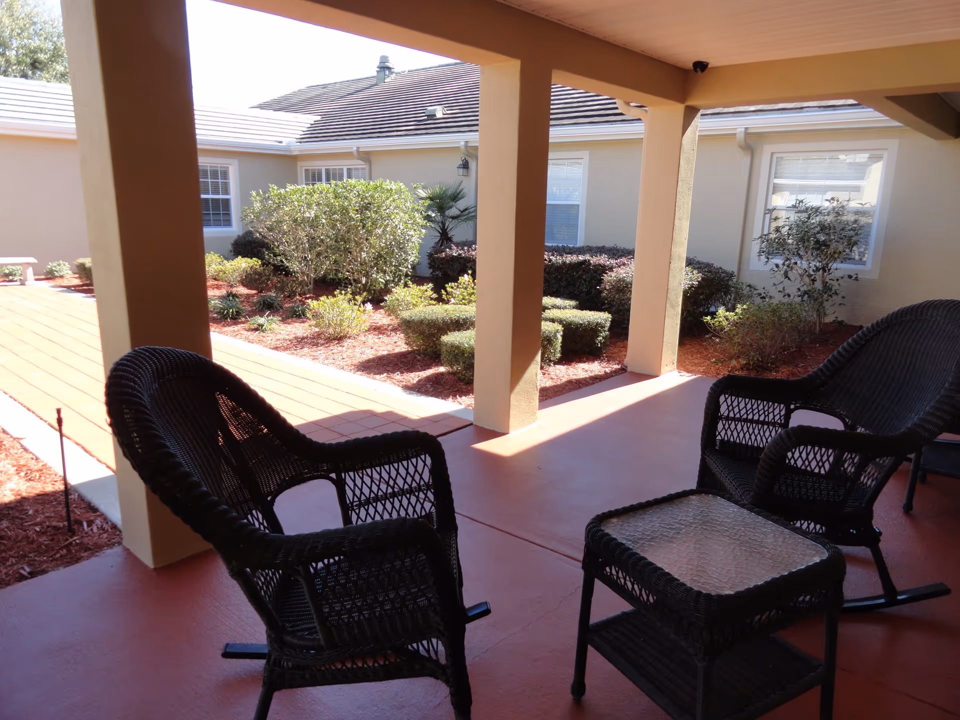 Covered outdoor patio area with two black wicker chairs and a matching glass-top wicker table, overlooking a landscaped garden with bushes and small trees in front of a beige building with white-framed windows.
