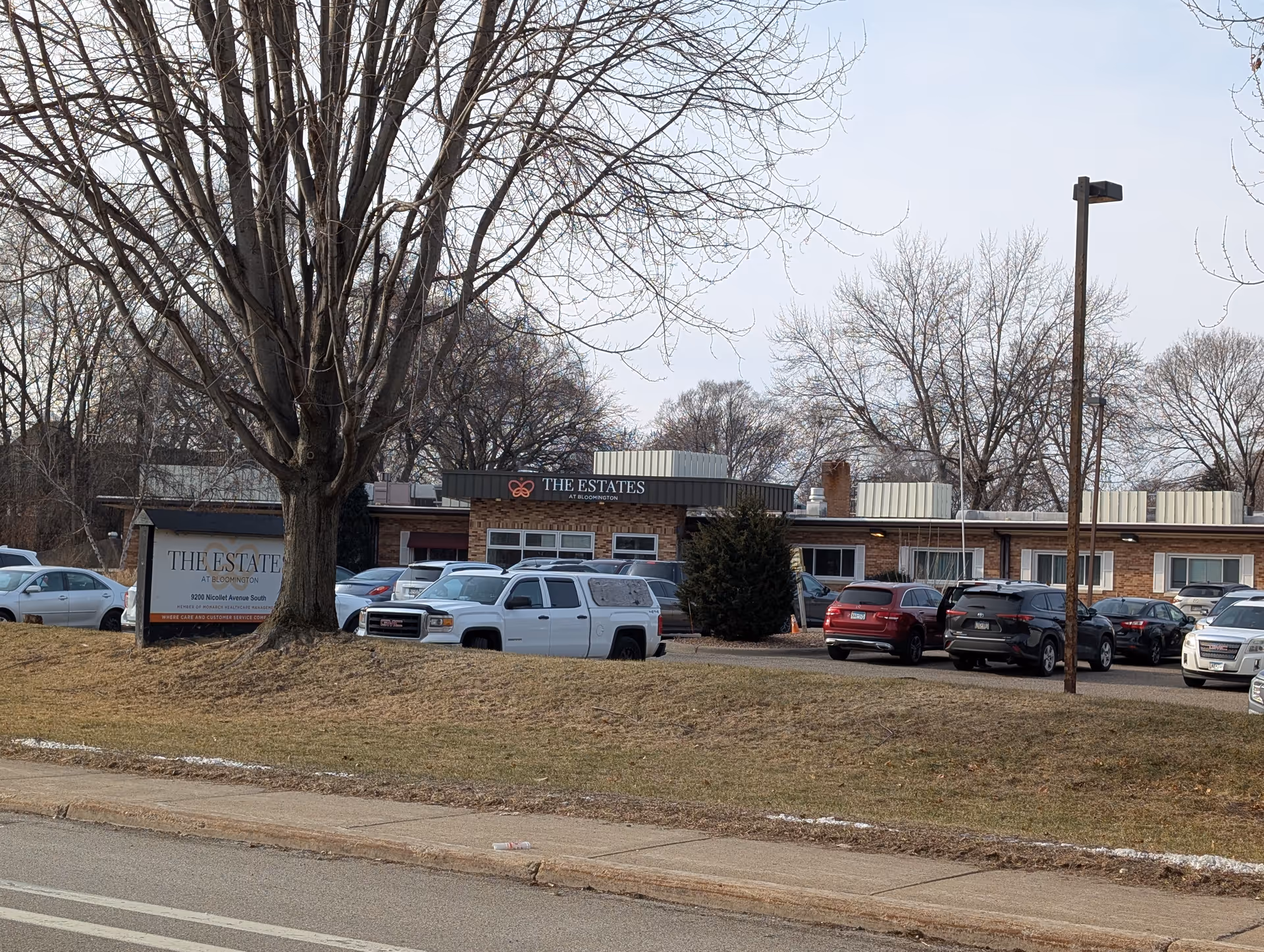 Front exterior of The Estates at Bloomington senior living building with a large tree and parked cars in the lot.