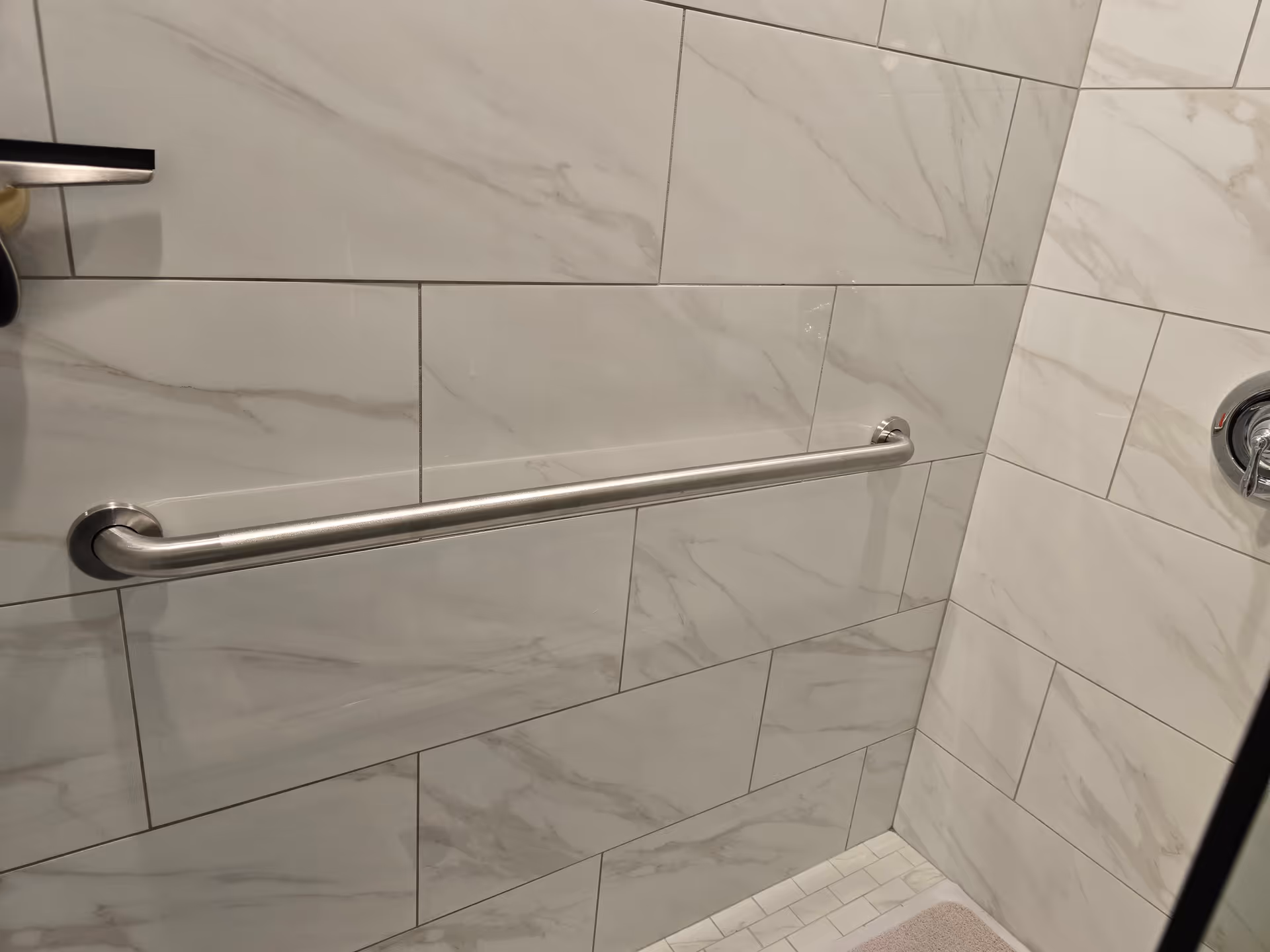 Shower area with a stainless grab bar mounted on marble-patterned tiled walls.