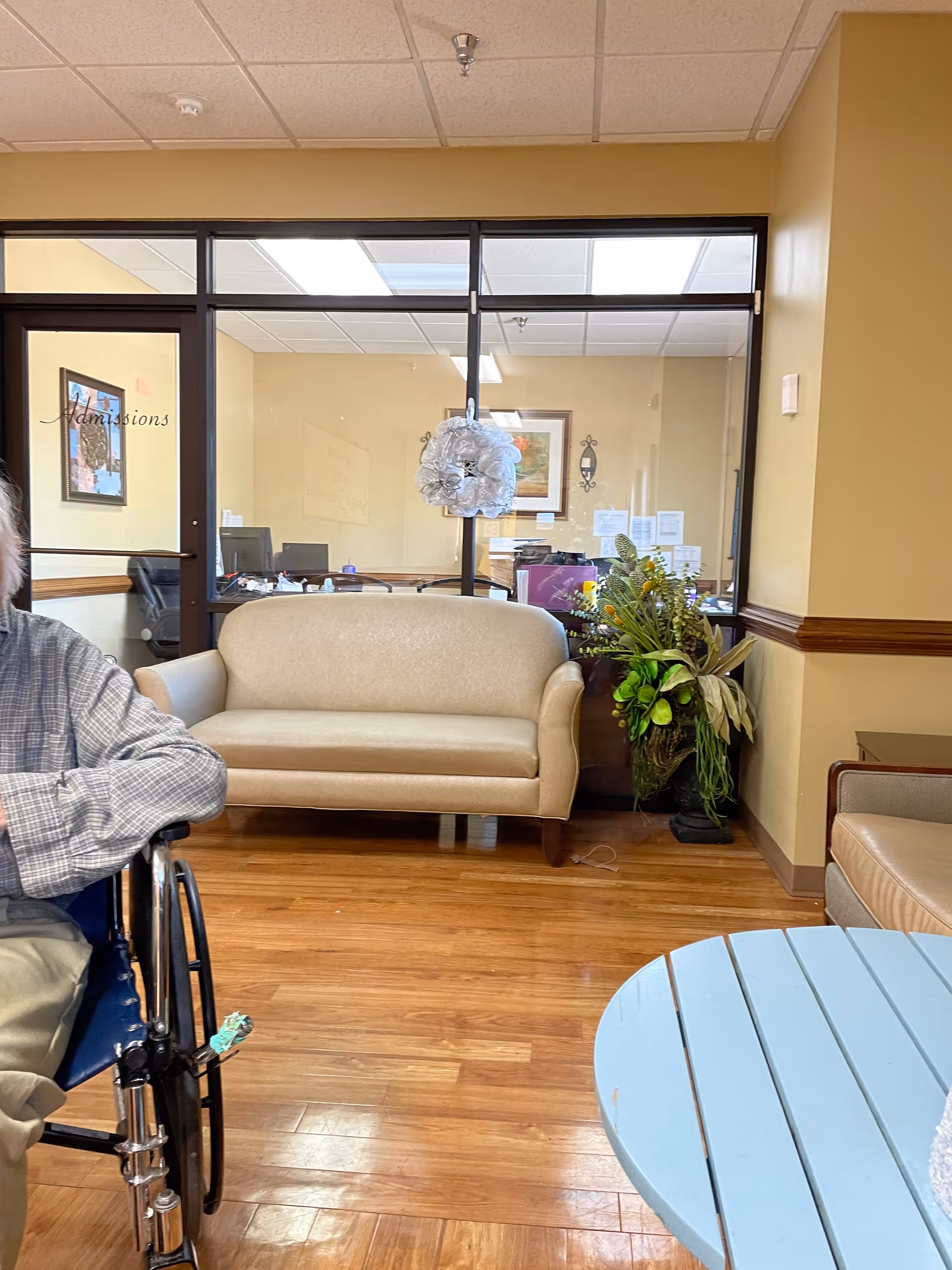 Interior lounge area with beige sofa, glass partition, wheelchair, potted plant, and blue round table.