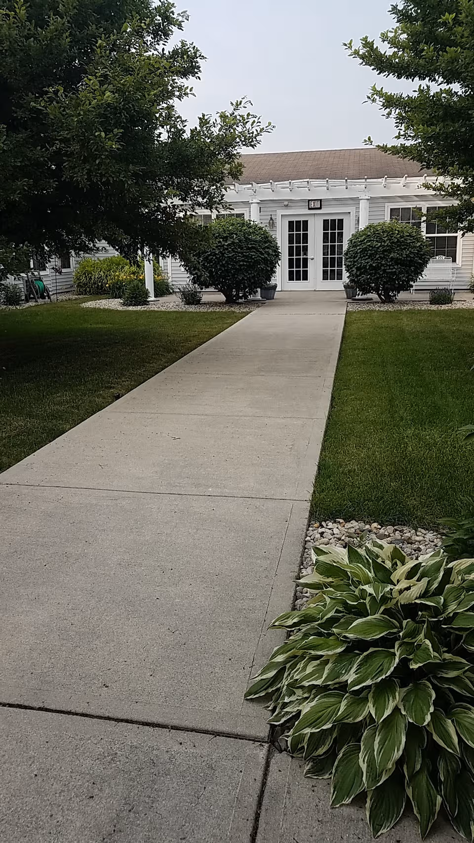 Concrete walkway leads to a white building entrance with double doors, flanked by trimmed bushes.