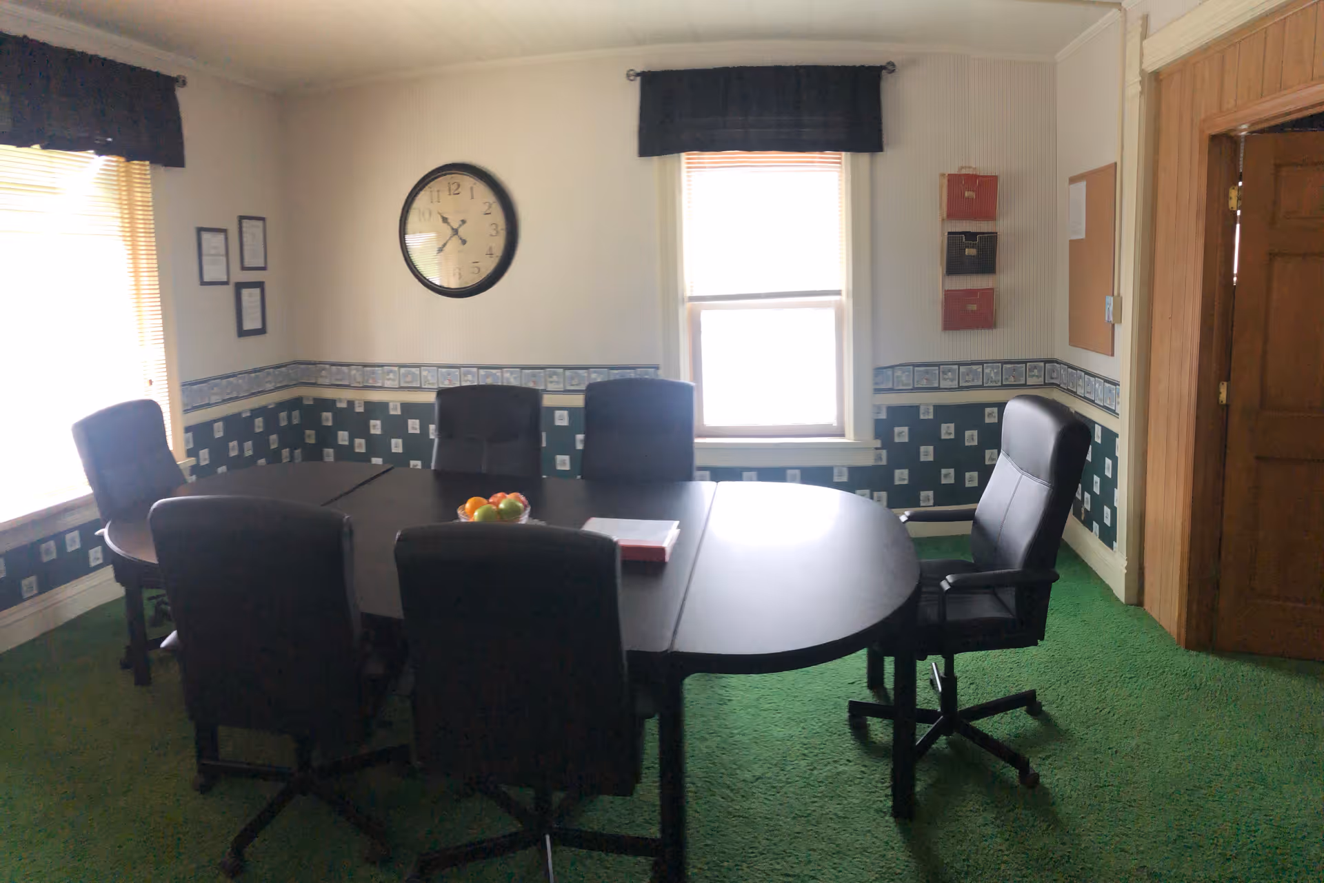 A conference-style meeting room with a large table, chairs, clock, and fruit bowl.