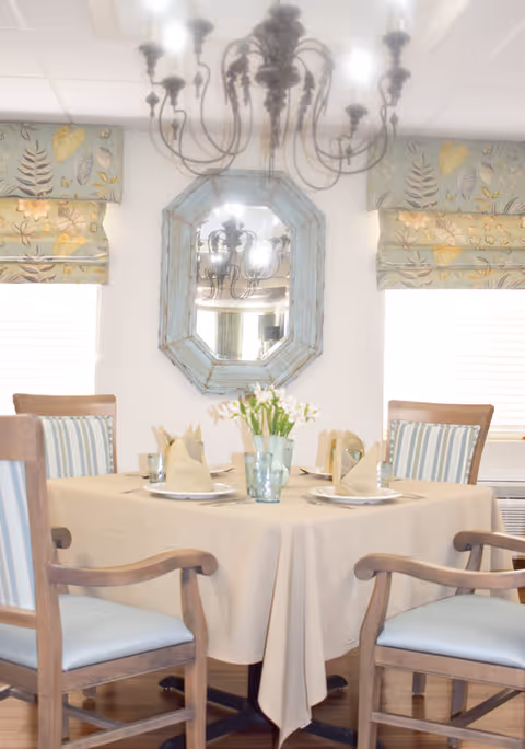 Round dining table set for four with folded napkins and a floral centerpiece beneath a chandelier in a bright dining room with a decorative mirror and upholstered chairs.