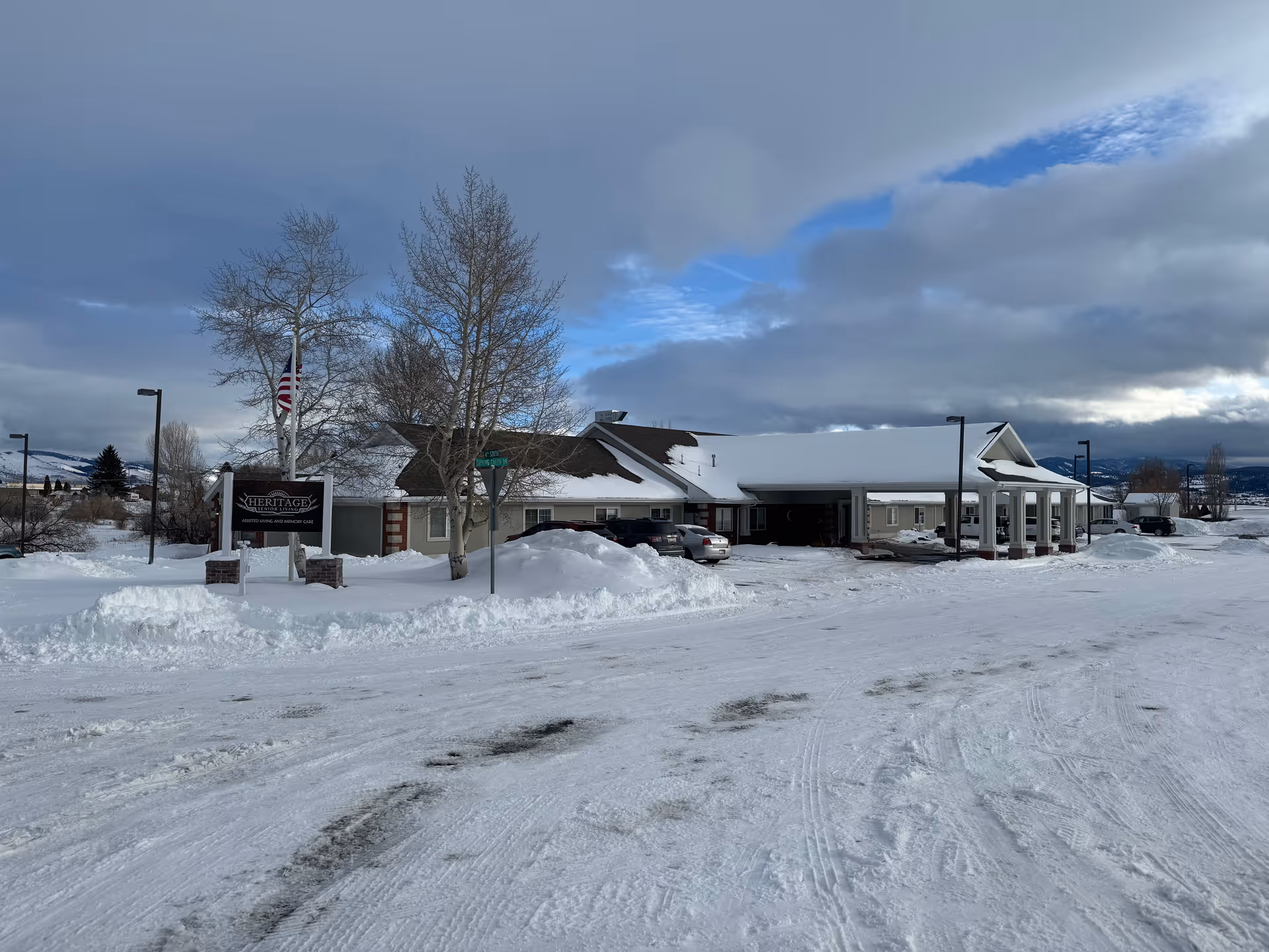 Snow-covered front exterior of a single-story senior living facility with an entrance canopy, parked cars, an American flag, and a Heritage sign under a cloudy sky.