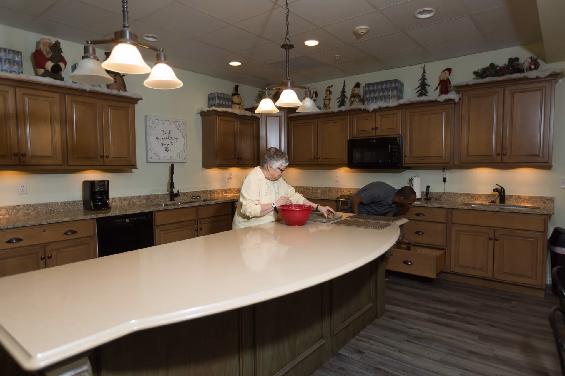 Senior living facility kitchen with a curved island, wood cabinets, and two residents cooking.
