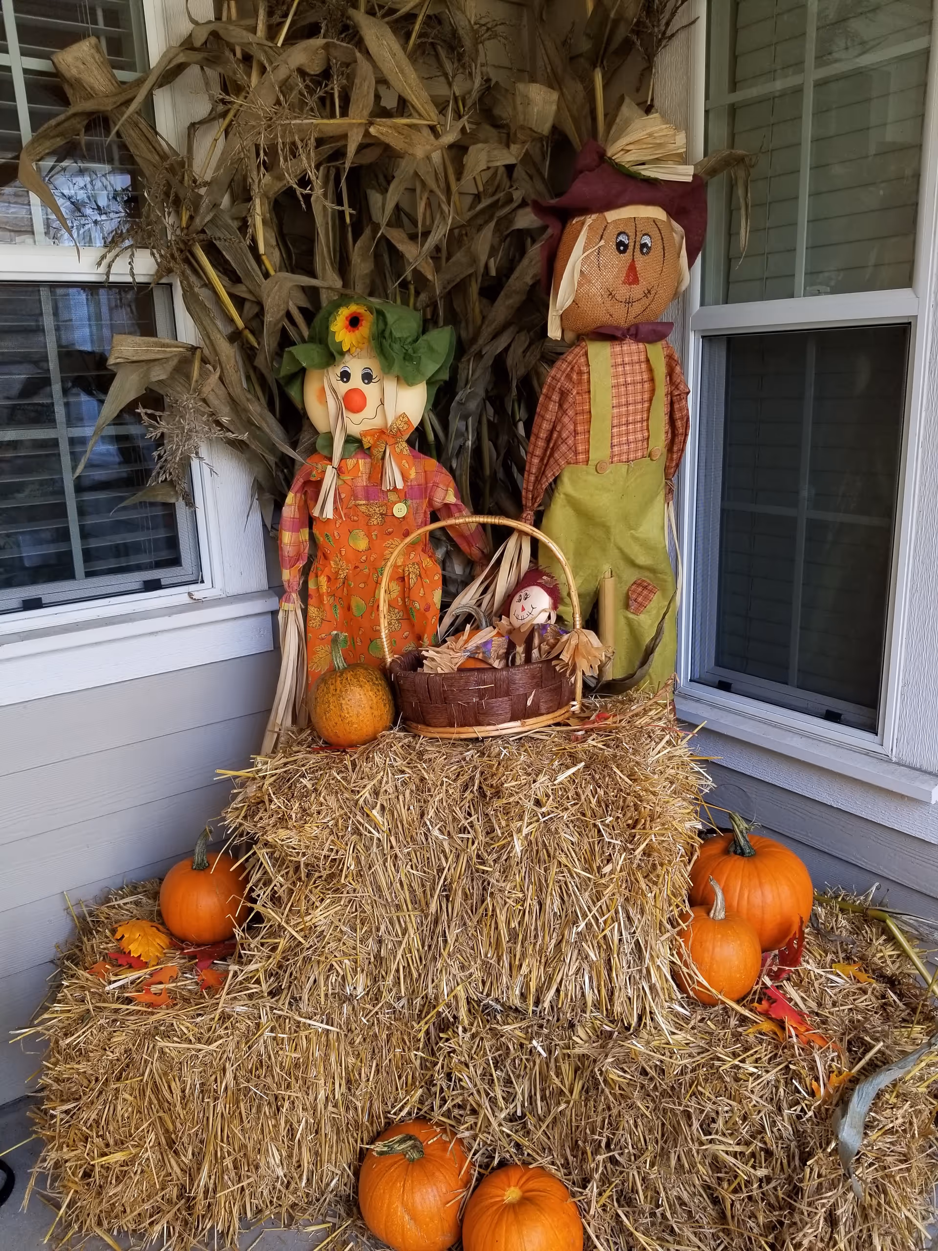 Outdoor fall display with hay bales, pumpkins, cornstalks, and scarecrow figures on a porch.