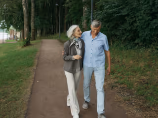 Two elderly people walk arm-in-arm along a park path outdoors.
