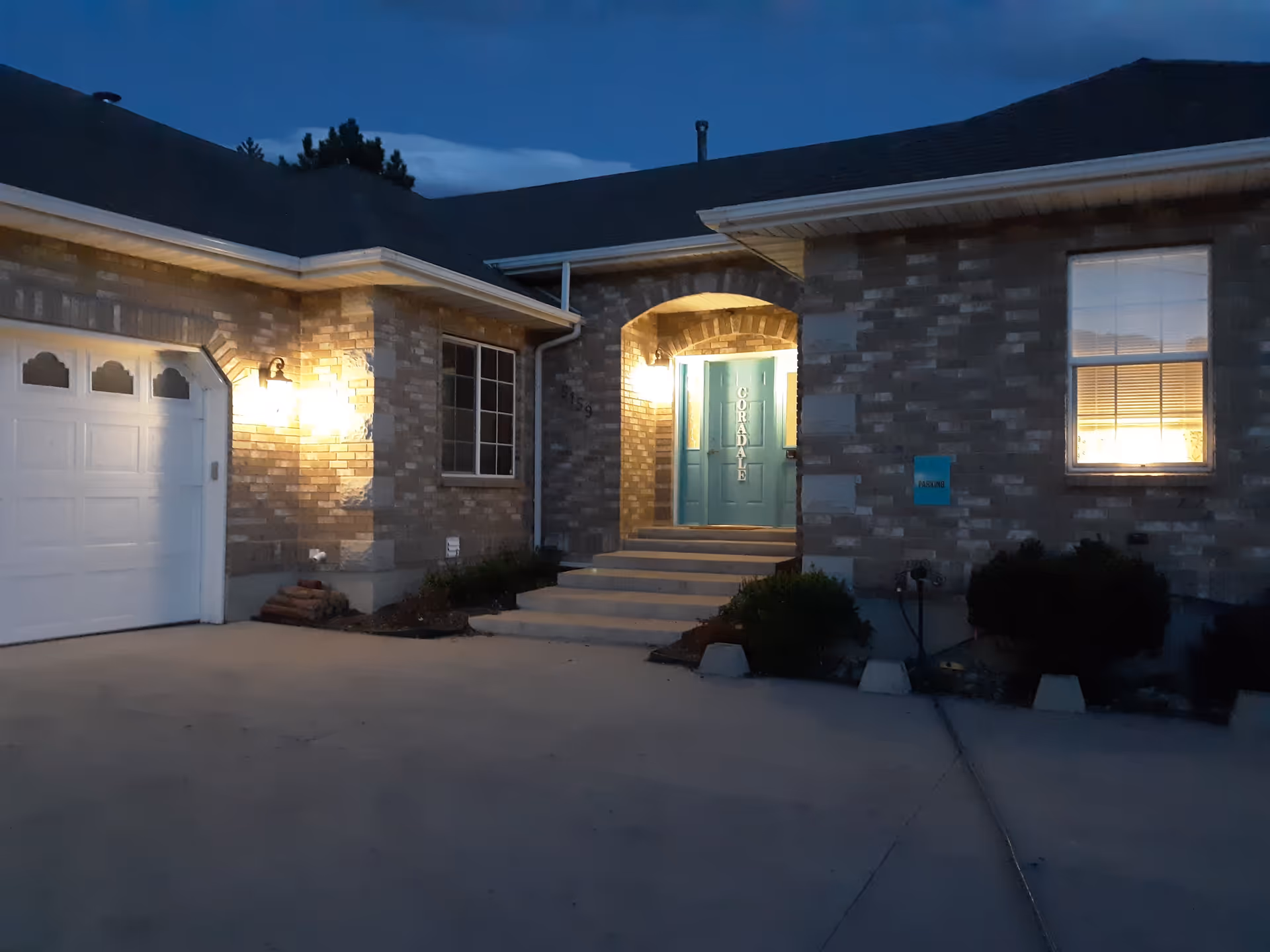 Exterior view of a brick residential building at dusk with a lit porch light. The entrance features a blue door with the word 'CORADALE' vertically displayed. There is a garage door on the left and a window with blinds on the right. The driveway and steps leading to the door are visible.