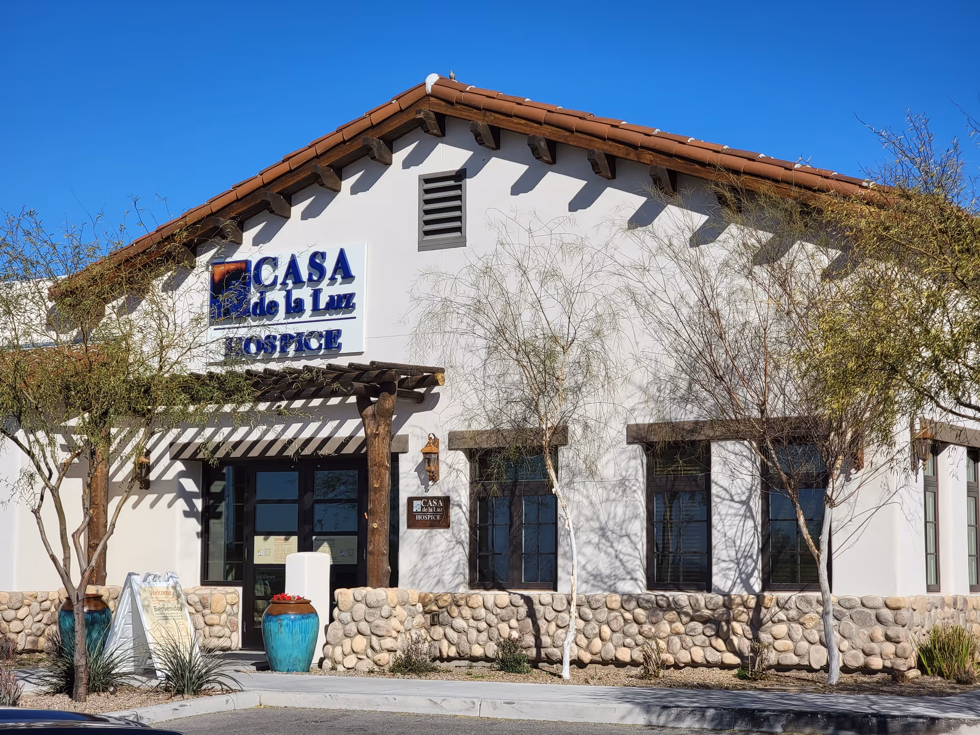 White hospice building with blue Casa de la Luz sign, stone base, and desert landscaping.