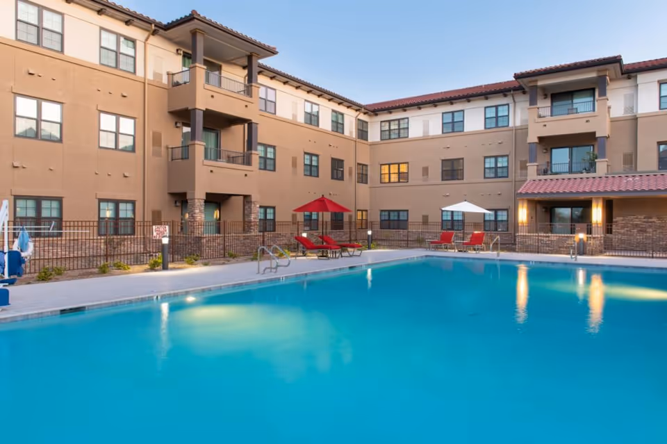 Outdoor swimming pool in a courtyard of a three-story senior living building with lounge chairs and umbrellas.