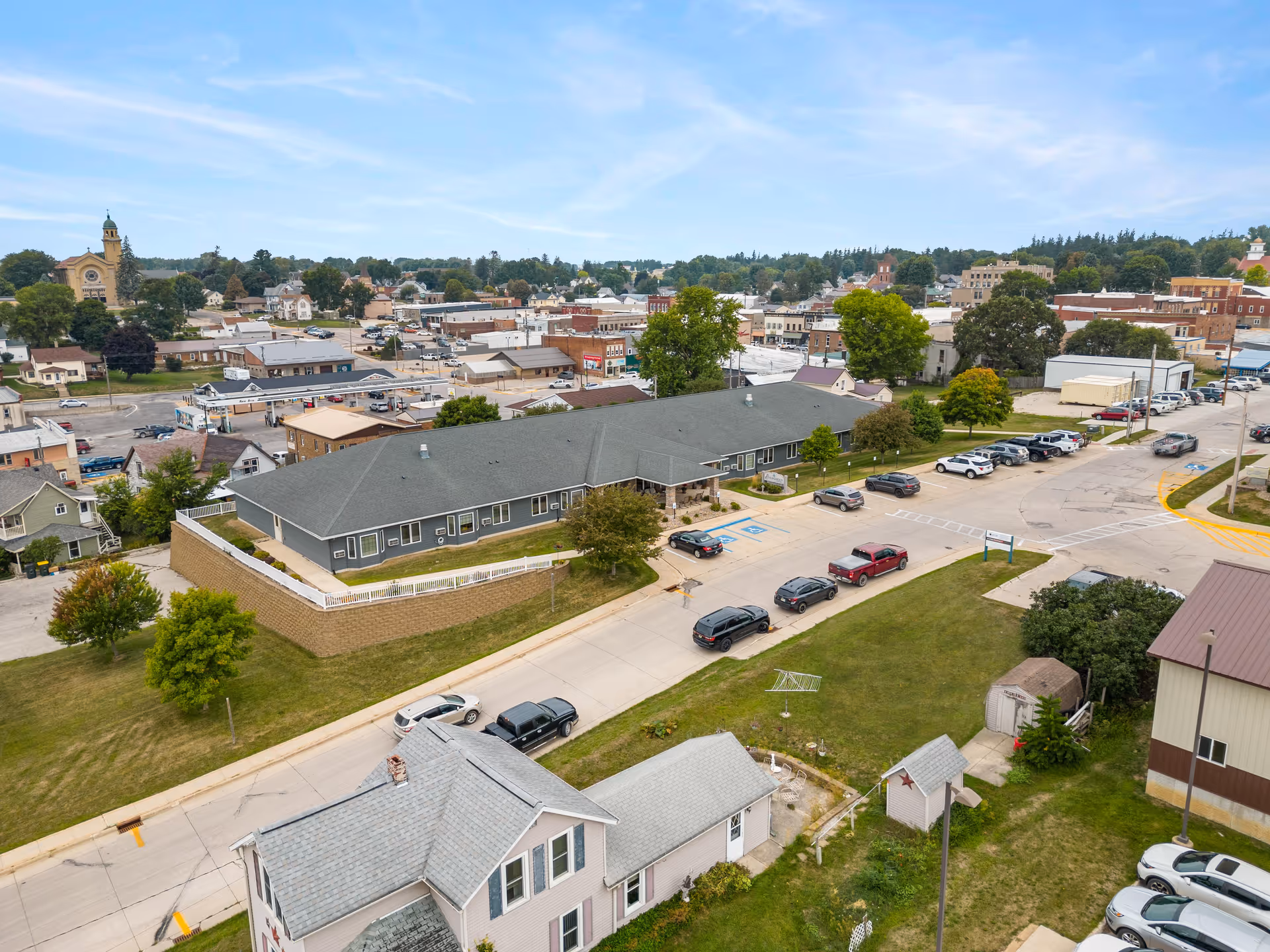 Aerial view of Good Samaritan Society - Waukon facility showing a large single-story building with a gray roof, surrounded by parking spaces with several cars parked. The building is situated in a small town with other houses, buildings, and trees visible in the background under a partly cloudy sky.