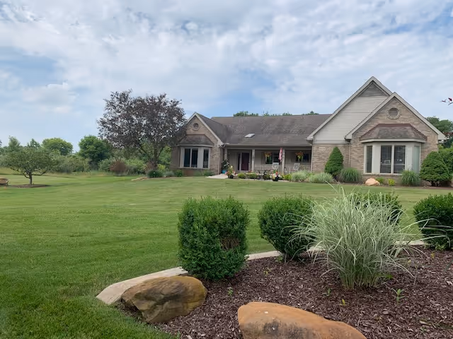 Single-story brick house with a large mowed front lawn and landscaped shrubs under a partly cloudy sky.