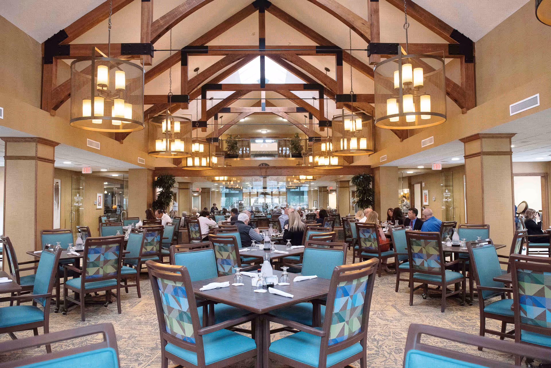 Spacious dining room in a retirement resort with multiple tables and chairs arranged neatly. The ceiling features exposed wooden beams and large hanging light fixtures. Several people are seated and dining in the background.