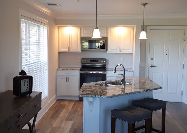 Open modern kitchen with a marble-topped island, stainless steel range and microwave, pendant lights, two stools, and an entry door.