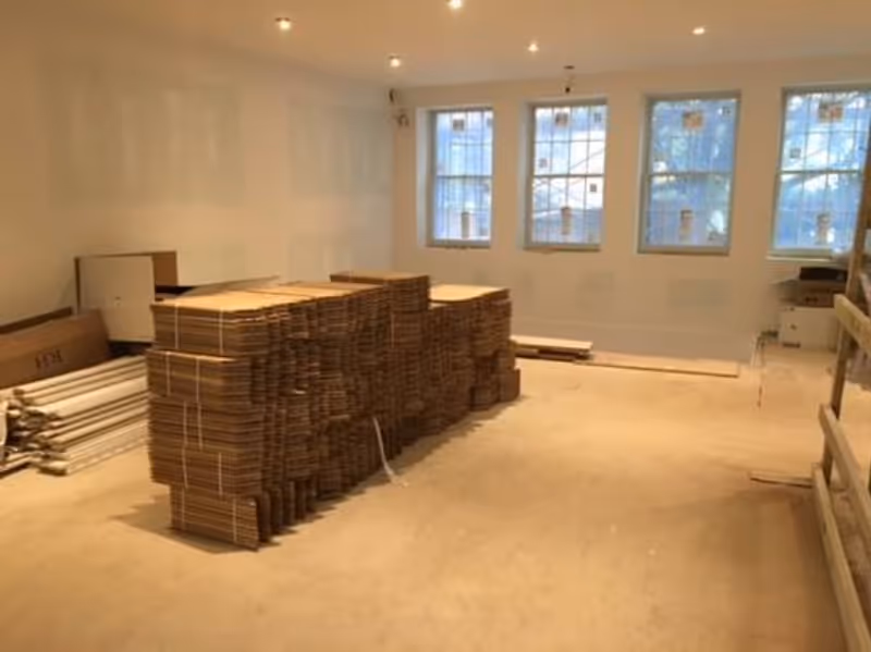 An unfinished indoor room with stacked plywood sheets and construction materials near three windows.