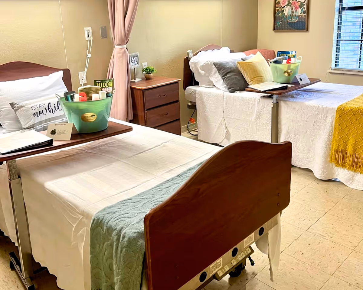 A nursing facility room with two hospital-style beds, each with white bedding and pillows. Each bed has a wooden headboard and a tray table holding a green basket with personal care items. Between the beds is a wooden nightstand with a small plant and framed photo. A window with blinds and a colorful painting are visible on the wall.
