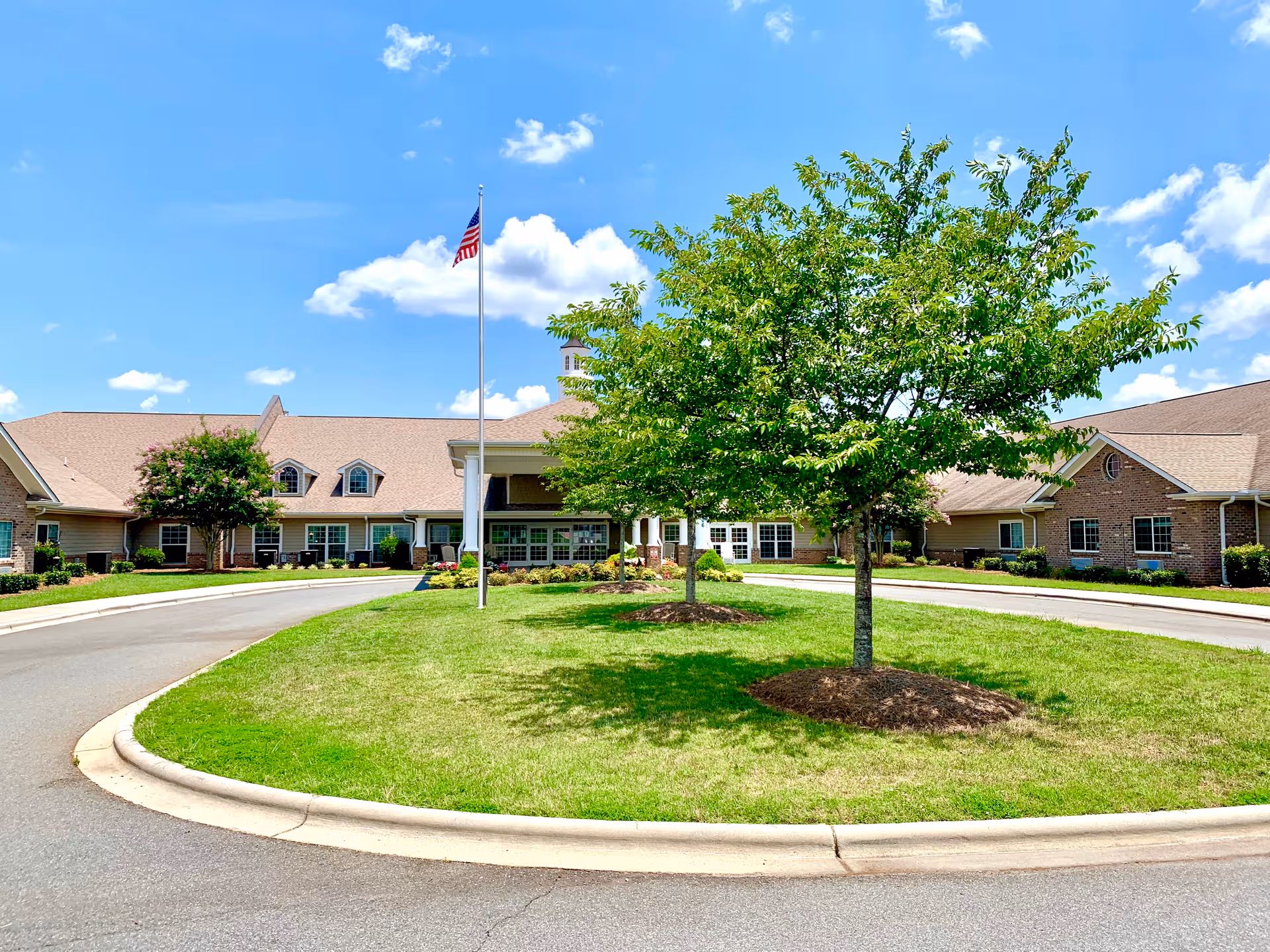 Exterior view of a senior living facility with circular driveway, flagpole, and trees.