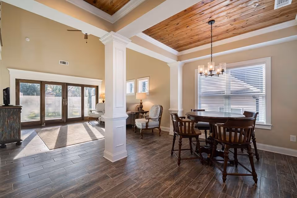 Bright open living and dining area with a wooden ceiling, round dining table and chairs, columns, and large glass doors letting in sunlight.