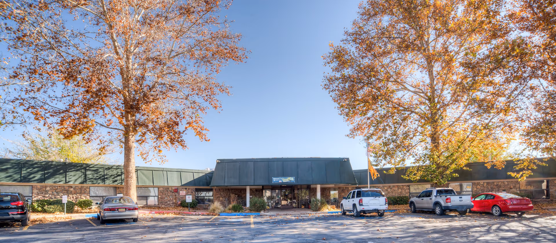 Front exterior view of Sandia Ridge Center, a single-story building with a green roof and stone facade, surrounded by large trees with autumn leaves and a parking lot with several parked cars.