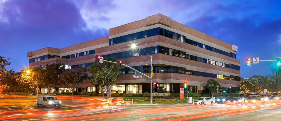Three-story brick senior living facility on a busy street at dusk with light trails.