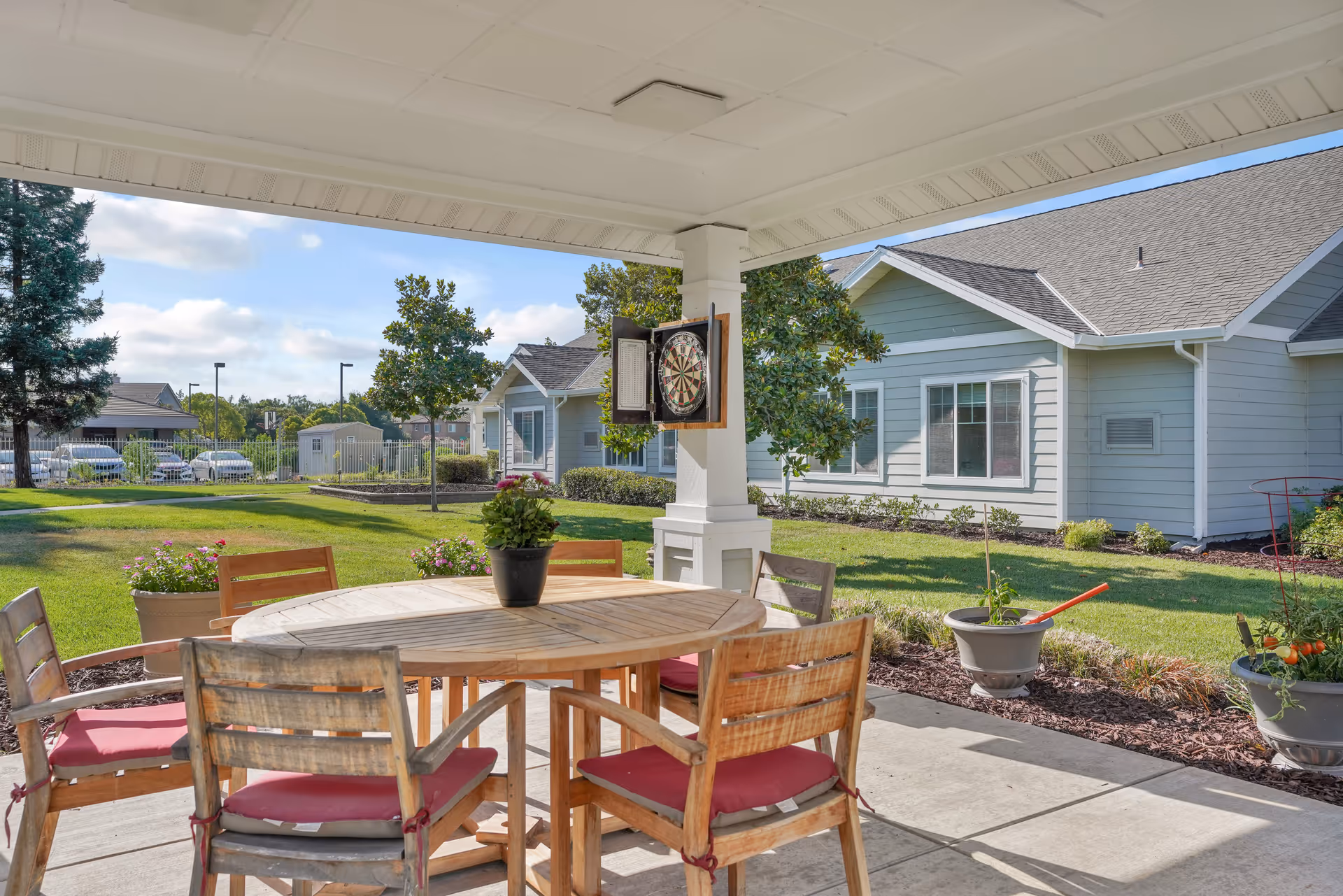 Covered outdoor patio area with a round wooden table surrounded by six wooden chairs with red cushions. A potted plant is on the table. A dartboard is mounted on a white pillar. In the background, there is a green lawn, flower pots, trees, and light blue residential buildings under a partly cloudy sky.