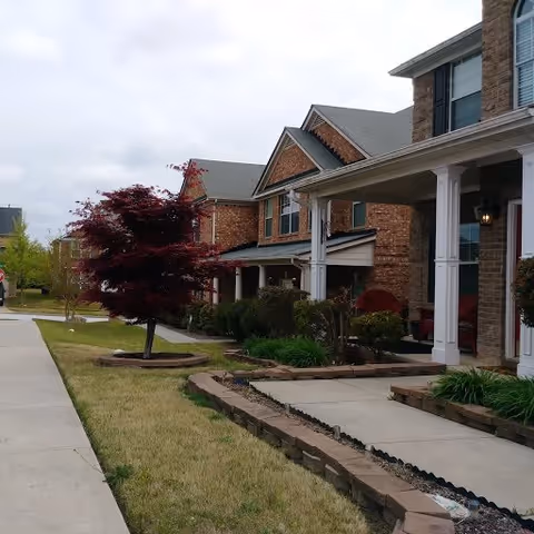 Exterior view of a brick residential building with a covered porch, landscaped garden beds, a small tree with red leaves, and a concrete walkway leading to the entrance.