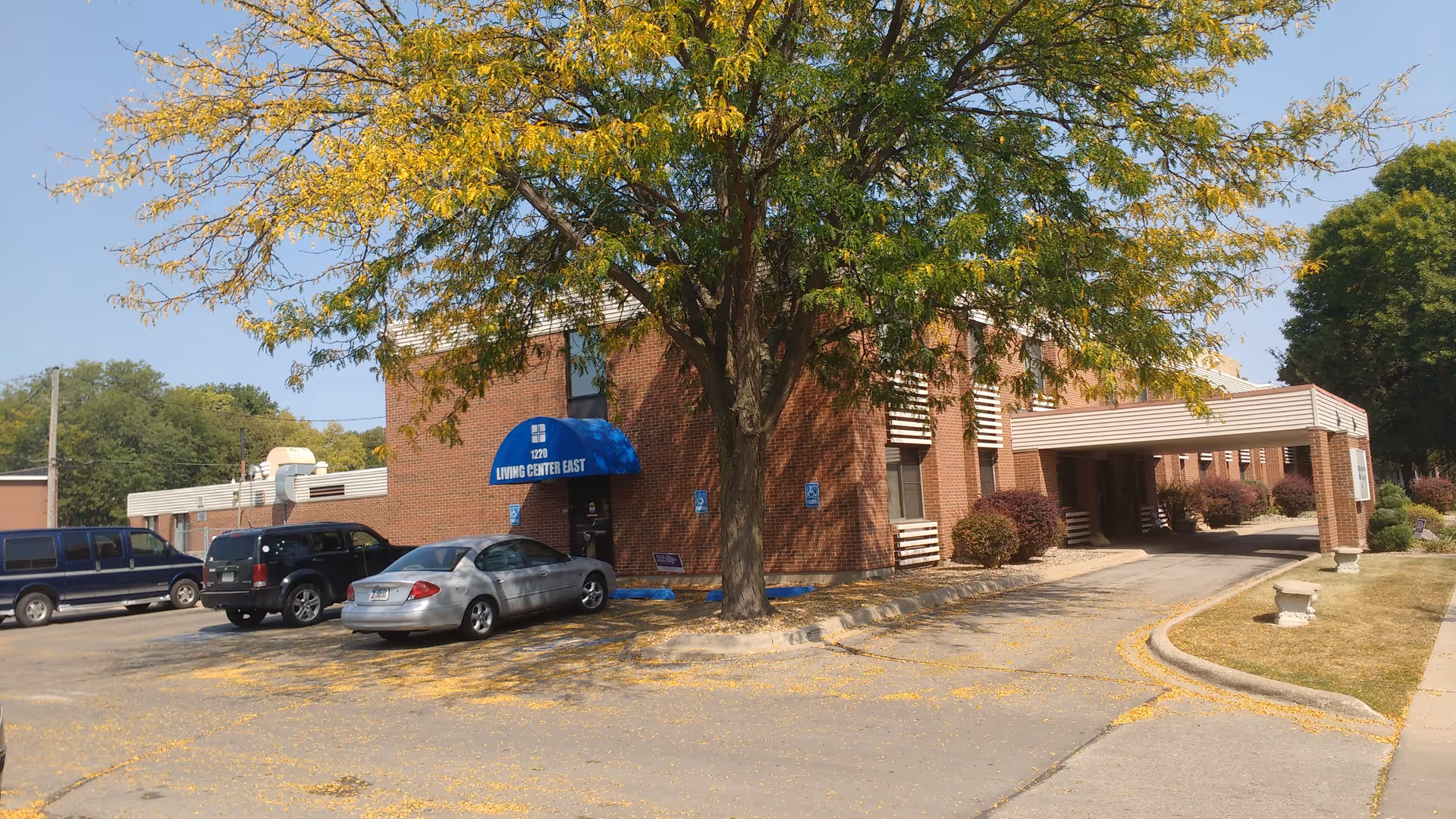 Brick senior living center exterior with parked cars and a large tree.