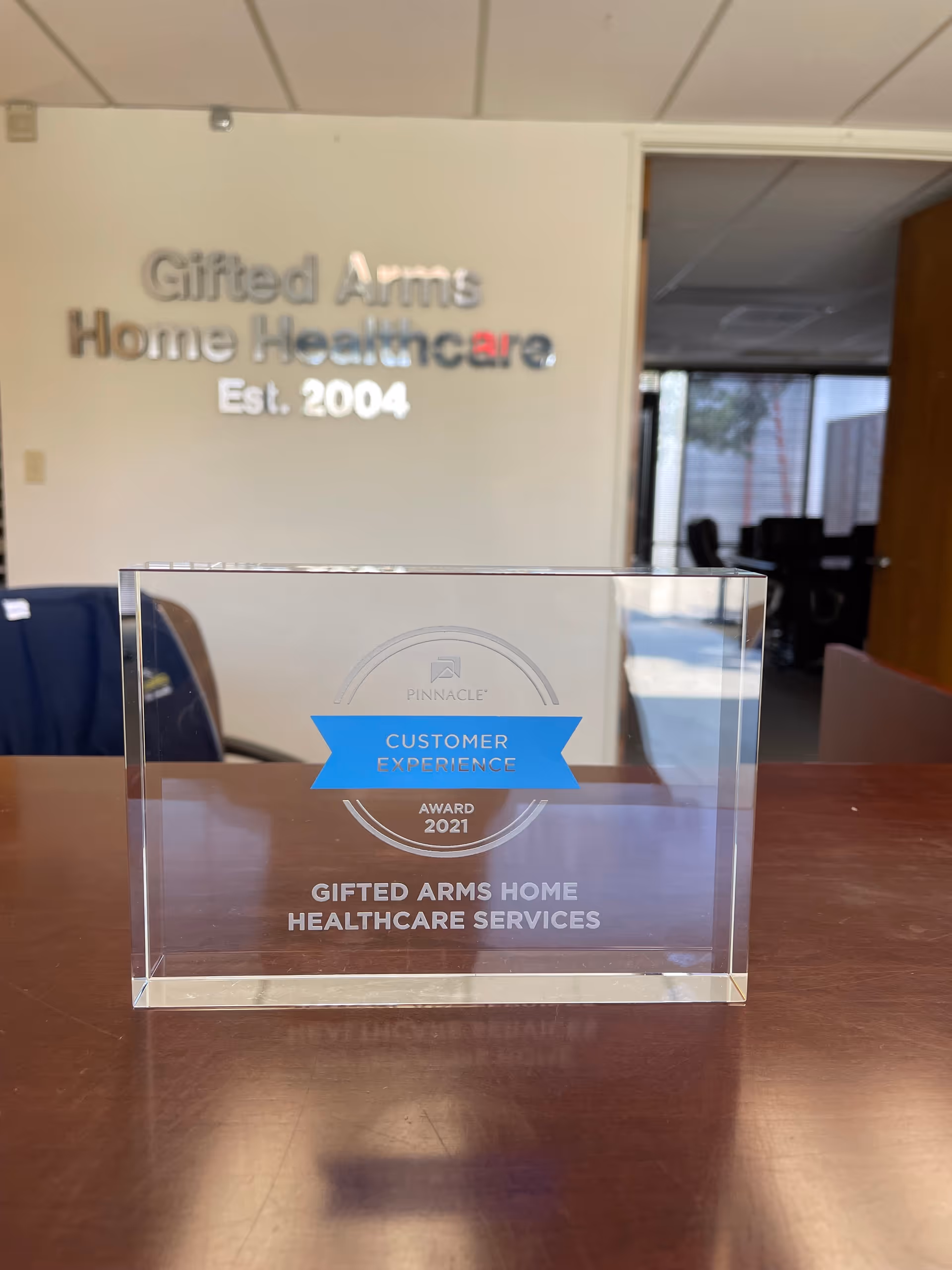 Clear acrylic award rests on a wooden desk with Gifted Arms Home Healthcare Services signage.