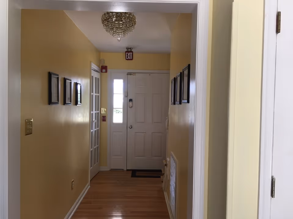 Yellow hallway with hardwood floor, framed pictures, and a chandelier in a senior living facility.