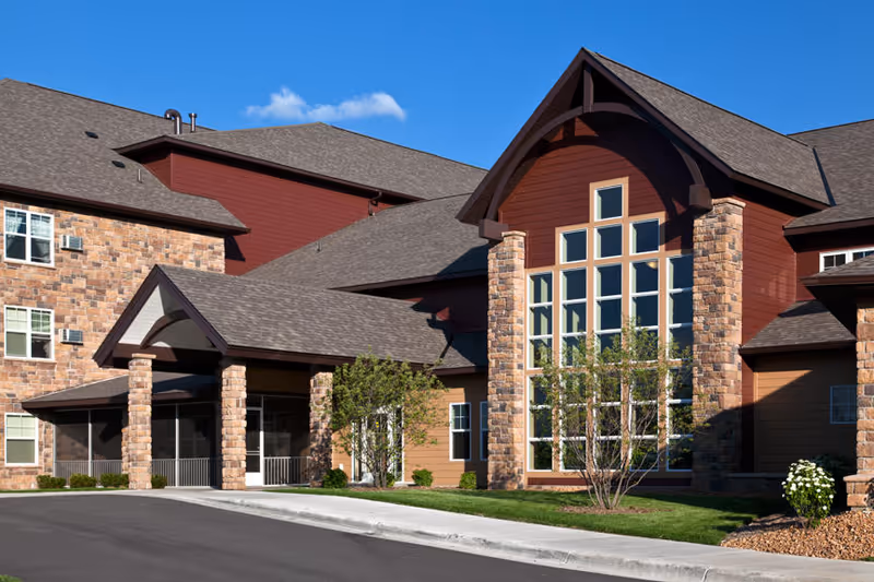 Front entrance of a multi-story senior living building with stone columns, a large arched window wall, covered drive-up, and landscaped lawn under a blue sky.