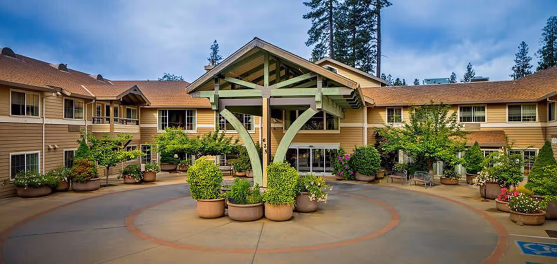 Front entrance of Eskaton Village Grass Valley, featuring a covered drop-off area with green wooden beams, surrounded by potted plants and shrubs, with a two-story building in the background under a partly cloudy sky.