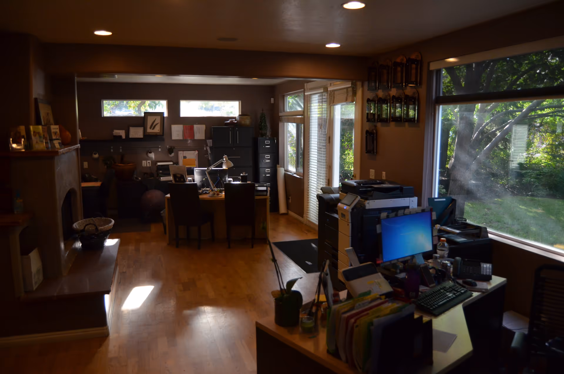 Dim-lit office/workspace with desks, computers, filing cabinets, and large windows overlooking greenery.