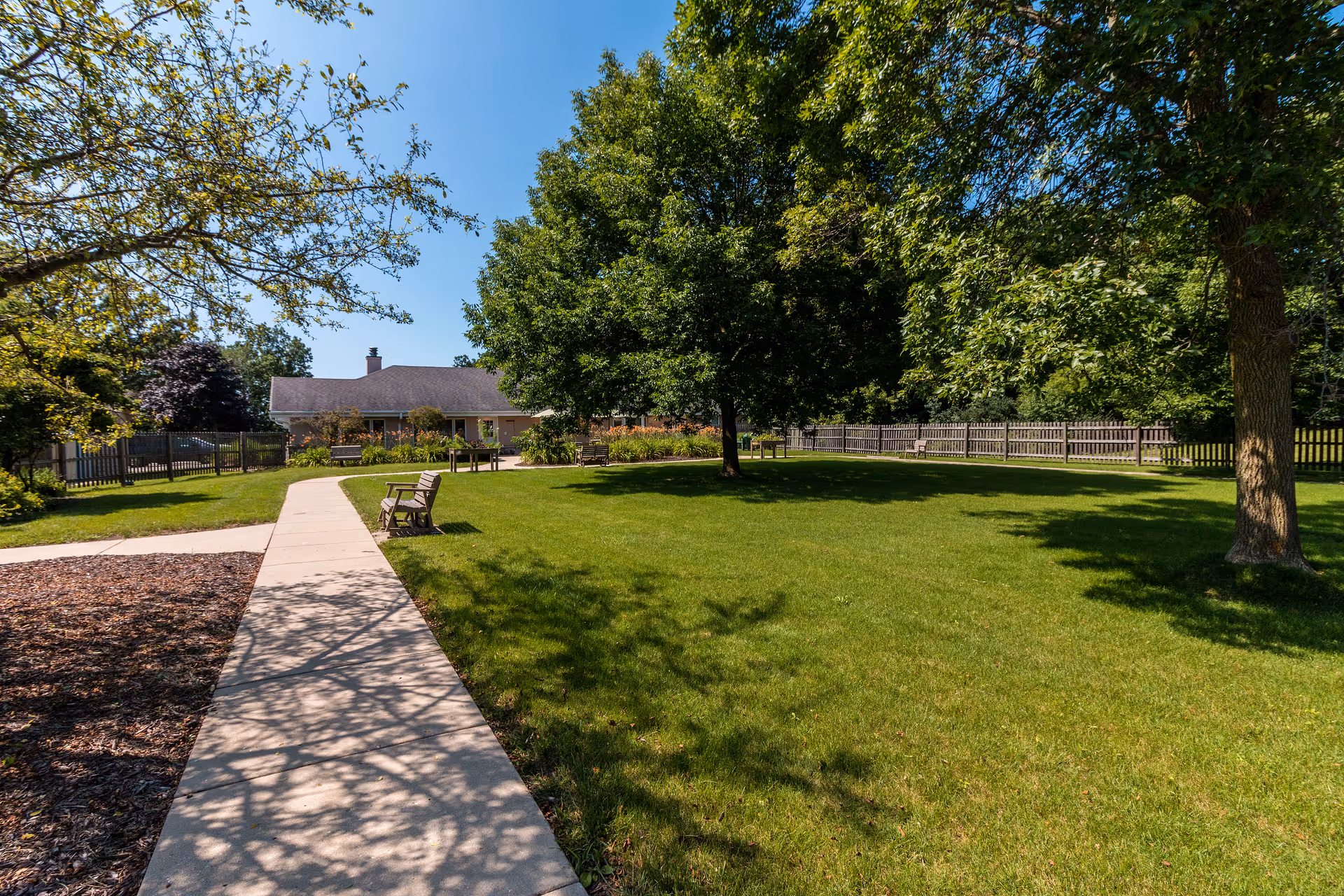 A sunny outdoor area at New Perspective Senior Living | Mequon featuring a concrete walkway leading towards a single-story building. The area is surrounded by green grass, several trees providing shade, wooden benches along the path, and a wooden fence enclosing the space.