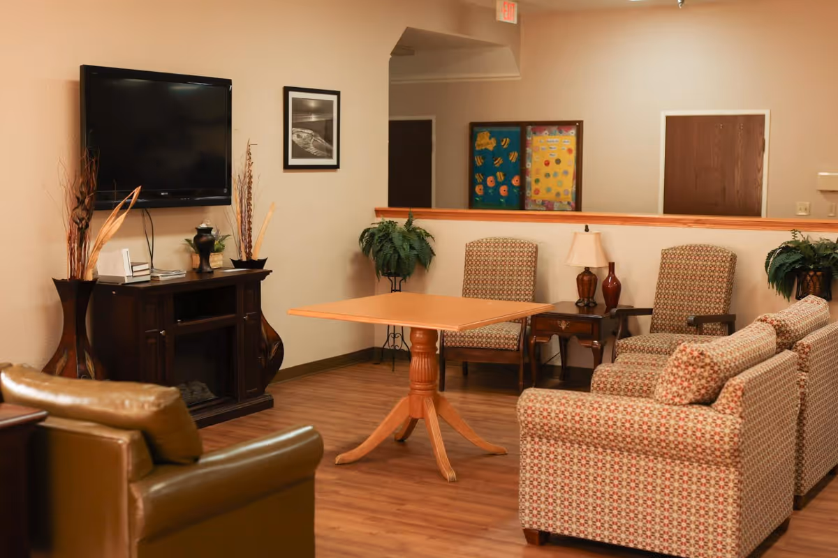 Common living room area with patterned armchairs, a central wooden table, wall-mounted TV, and decorative plants.