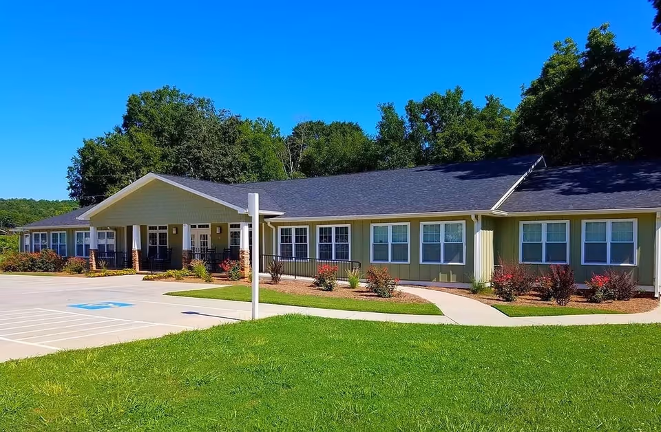 Single-story senior living building with a covered entrance, parking spaces, and landscaped lawn under a clear blue sky.