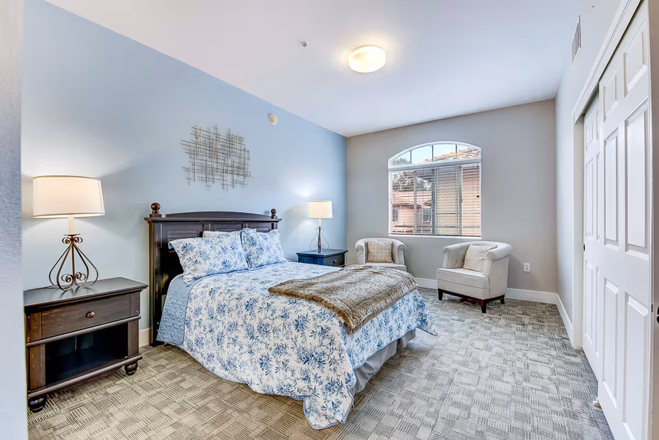 A furnished bedroom featuring a queen bed, two nightstands, lamps, and window seating.