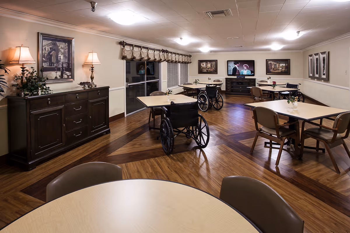 A well-lit dining room in a senior living facility with several tables and chairs, including some wheelchairs. The room features wood-patterned flooring, a sideboard with lamps and a framed picture, large windows with valances, and a television mounted on the far wall showing a scene from a movie or show. There are also framed pictures on the walls.