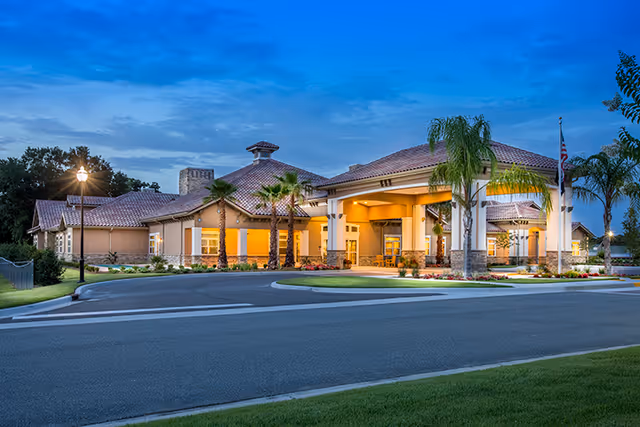 Front entrance of a well-lit single-story assisted living facility with palm trees and a circular driveway at dusk.