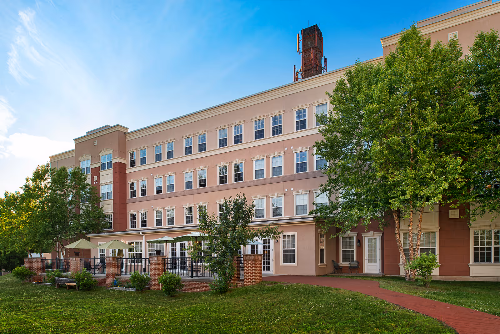 Exterior view of a multi-story senior living facility building with multiple windows, a patio area with umbrellas and seating, surrounded by green trees and a grassy lawn under a blue sky.