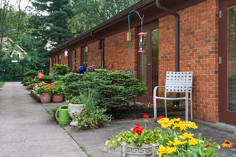 Outdoor walkway alongside a brick building with multiple doors and windows. The walkway is lined with various potted plants and flowers, a green watering can, and a white chair. There are bird feeders hanging near the doors and lush green trees in the background.