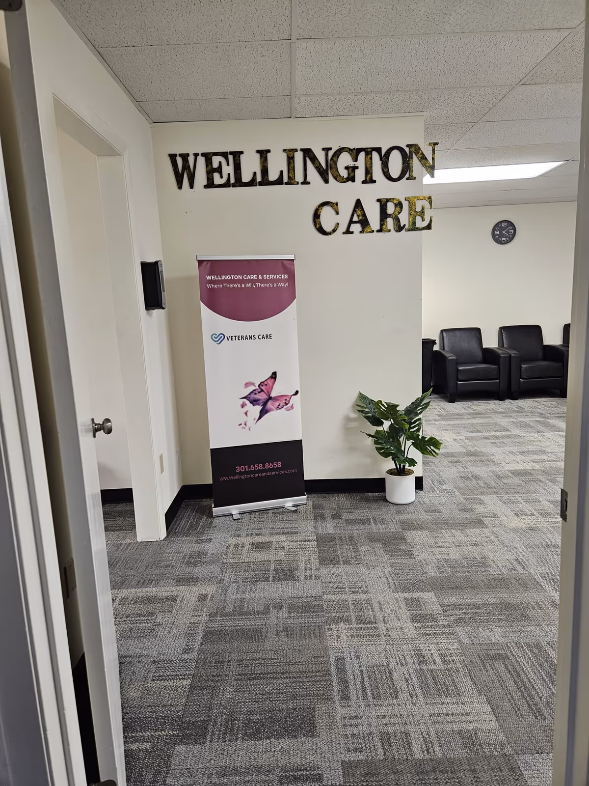 Interior hallway of Wellington Care with sign, banner, plant, and waiting chairs.