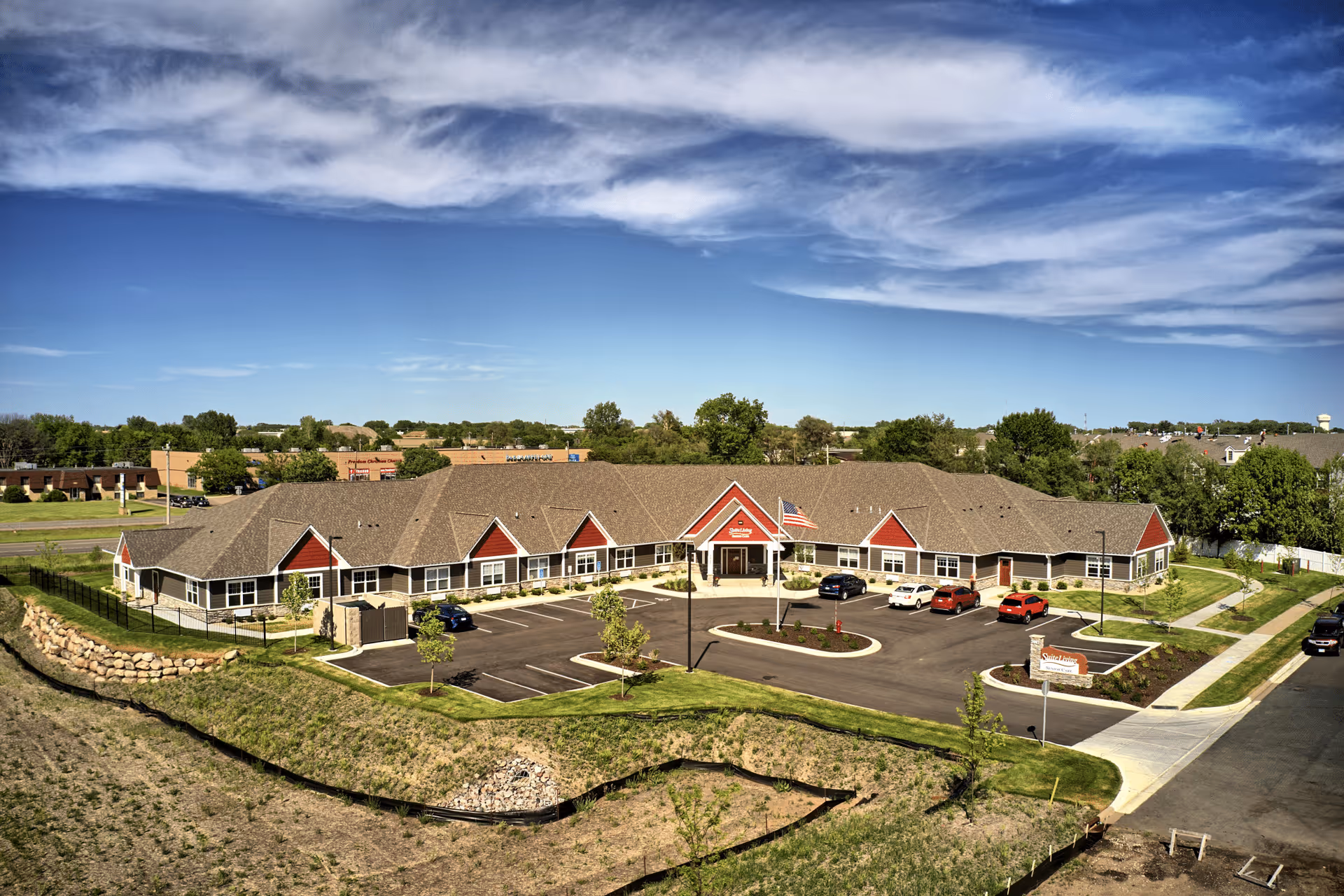 Aerial view of Suite Living Memory Care & Assisted Living facility showing a single-story building with a brown roof and red accents. The building is surrounded by a parking lot with several cars, landscaped greenery, and a clear blue sky with some clouds above.